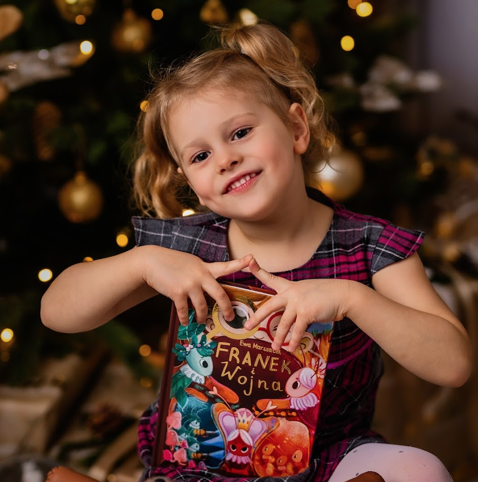 Pięcioletnia dziewczynka trzyma w rękach książkę dla dzieci "Franek i Wojna"